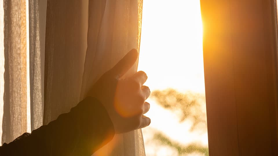 A hand reaching to open a curtain with the sun shining through the gap